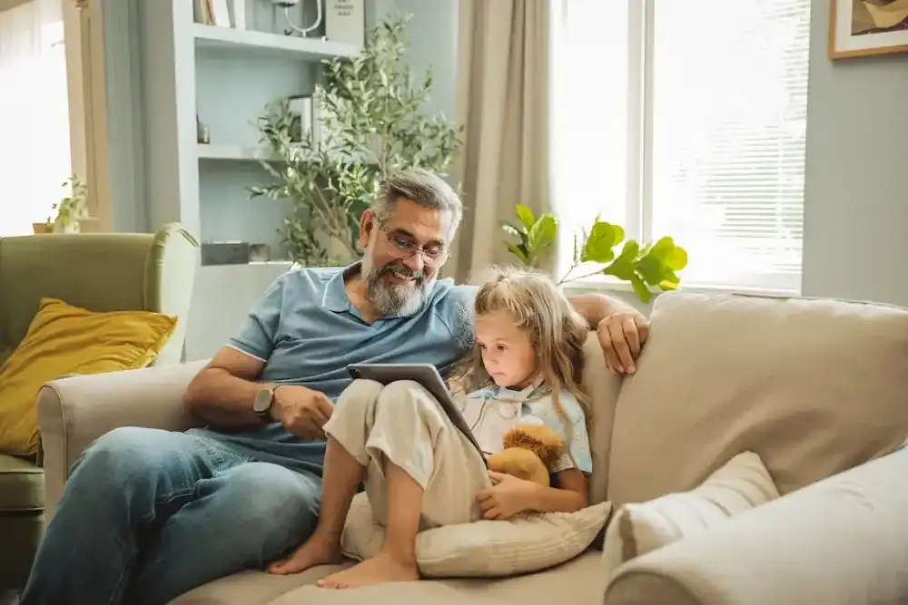 A smiling grandfather and his young granddaughter sitting together on a sofa while looking at a tablet.