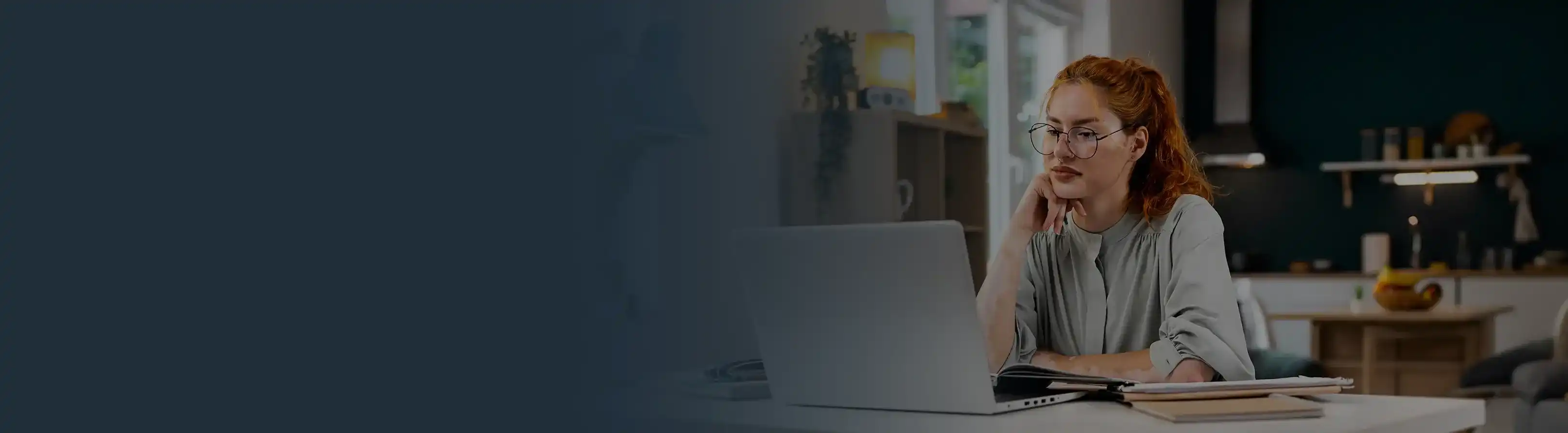 A young woman with glasses sitting at a white desk and working on a laptop at home
