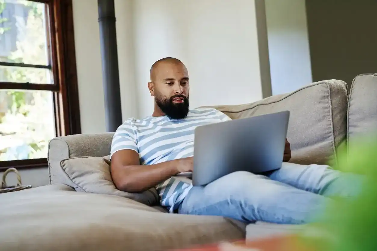 A man with a beard wearing a striped shirt relaxes on a grey sofa while using a laptop computer.